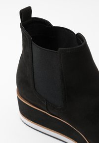 Czarne botki za kostkę z zamszu z teksturowanym elastycznym panelem. Posiadają grubą podeszwę na koturnie oraz kontrastowe akcenty w białym i brązowym kolorze.