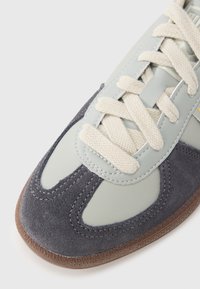 Tênis com parte superior em couro verde claro com detalhes em camurça cinza, atacadores creme e sola de borracha texturizada castanha. Acabamento liso e durável.