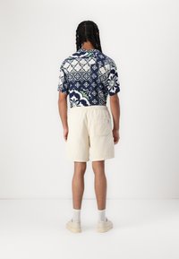 Kurzärmliges marineblaues Hemd mit floralem Muster, kombiniert mit cremefarbenen Shorts. Das Hemd zeichnet sich durch komplexe Designs mit grünen und weißen Akzenten aus.