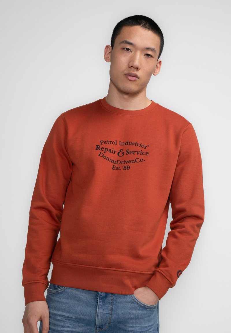 Felpa color ruggine in cotone, con scollatura rotonda e polsini a coste. Grafica frontale: "Petrol Industries Riparazione & Servizio."