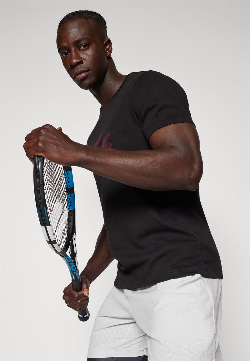 Chemise noire à manches courtes, shorts gris avec un motif texturé. La personne tient une raquette de tennis avec un accent bleu et une prise noire. Fond neutre.