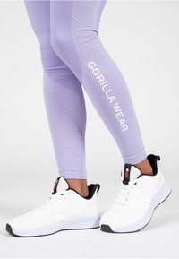 Paarse leggings met een textuurpatroon en witte "GORILLA WEAR" branding. Gecombineerd met witte sportschoenen met zwarte accenten.