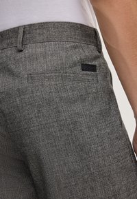 Primo piano di una persona che indossa pantaloni grigi con motivo a spina di pesce, dotati di tasca posteriore a filetto e etichetta nera del marchio Joop!