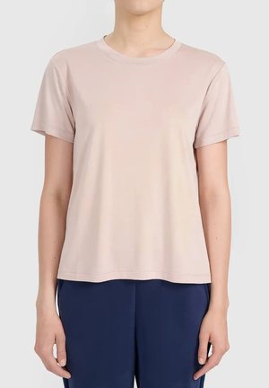 Personne portant un t-shirt rose clair uni à manches courtes et un pantalon bleu foncé, debout devant un fond blanc uni.
