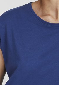 Close-up van een persoon die een eenvoudige blauwe ronde hals t-shirt draagt, met zicht op de schouder, hals en een deel van de bovenarm.