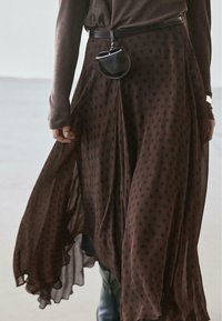 Brown polka dot pleated skirt with a flowing design, paired with a black leather belt bag accentuating the waist. Textured fabric.