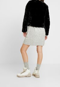 Kvinde iført en sort faux pelsjakke, grå strikket miniskirt, grå ankelstrømper og hvide vandrestøvler, stående foran en ensfarvet baggrund.