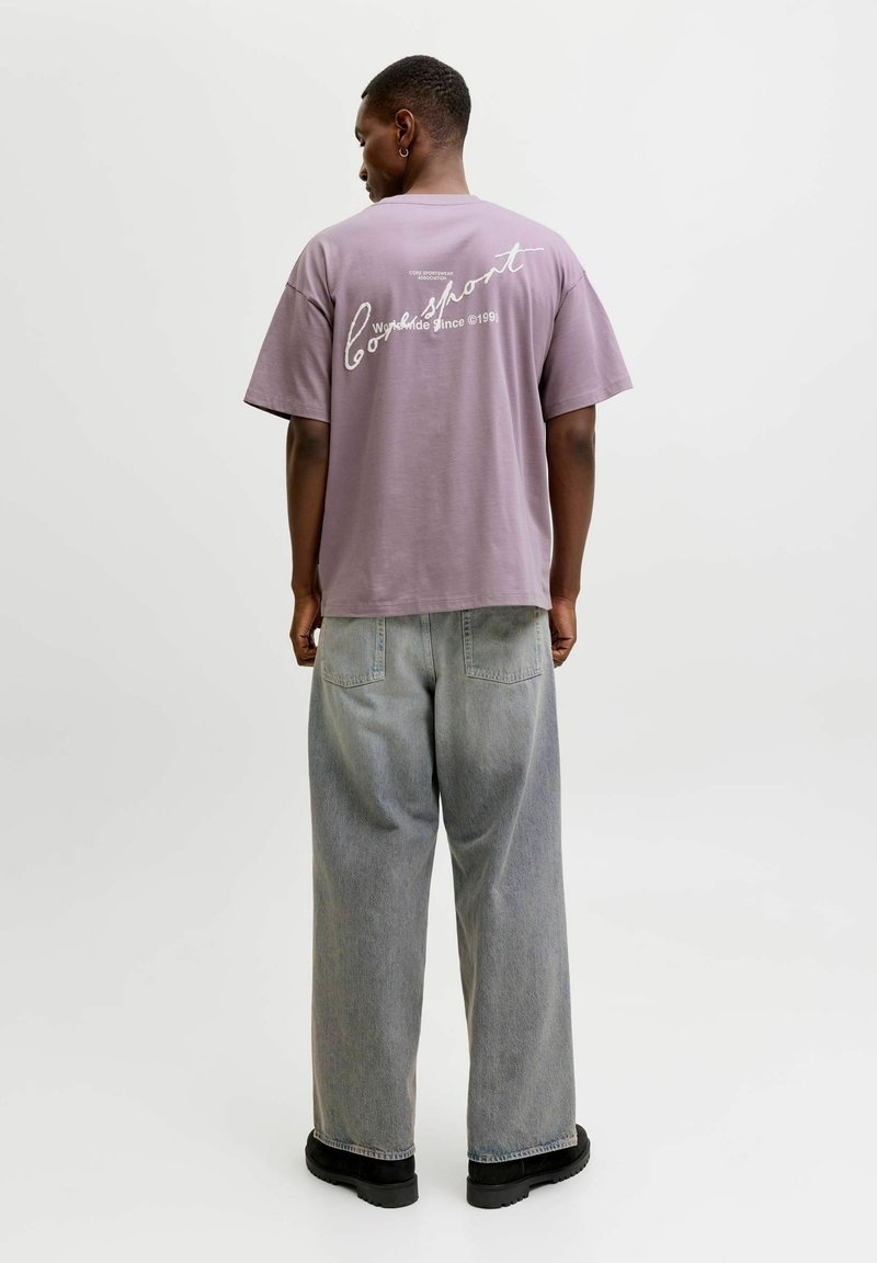 Vyras, vilkintis laisvo stiliaus purpurinės spalvos marškinėlį su baltais užrašais nugaroje, šviesiai pilkus platėjančius džinsus ir juodus batus, stovintis atsisukęs.