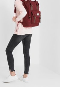 Bordowy plecak z materiałowym wykończeniem, dwoma górnymi uchwytami, skórzanymi akcentami, przednią kieszenią i regulowanymi paskami, noszony z czarnymi dżinsami i białymi trampkami.