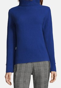 Kvinna iklädd en åtsittande kungligt blå polotröja och grå rutiga byxor med avslappnade händer vid sidan mot en vit bakgrund.