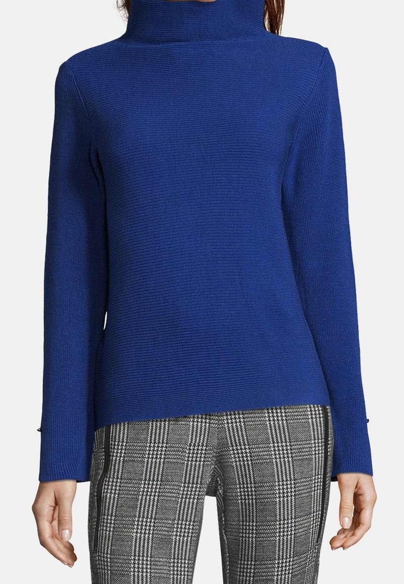 Kvinna iklädd en åtsittande kungligt blå polotröja och grå rutiga byxor med avslappnade händer vid sidan mot en vit bakgrund.