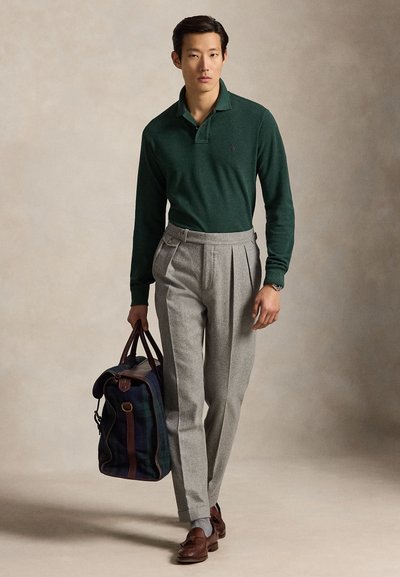 Man draagt een donkergroene lange mouwen polo, lichtgrijze geplooide broek, bruine loafers, draagt een donker geruite leren reistas.