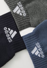 Trys poros kojinių pilkos, tamsiai mėlynos ir mėlynos spalvų, su raukšlėta tekstūra ir balta siuvinėta logotipu ant kiekvienos.