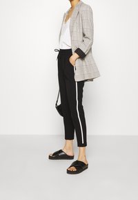 Svarta byxor med vita sidosträck, kombinerade med en grå rutig kavaj. Svarta slidesandaler fulländar stilen. Enkel, skräddarsydd design.