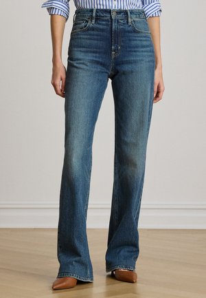 Personne portant un jean bleu taille haute et une chemise rayée, debout sur un sol en bois avec des chaussures marron pointues visibles.