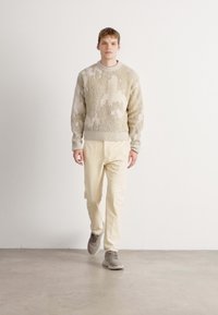 Jonge man die binnen loopt in een beige gestructureerde trui, crèmekleurige broek en grijze sneakers tegen een effen witte muur.