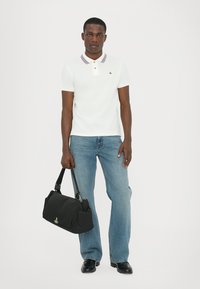 Balts polo krekls ar purpura svītroto apkakli, oranžu pogu, zila džinsi un melna auduma sporta soma ar logo, uz viendabīga fona.