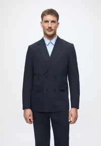 Dunkelblaues, doppelt geknöpftes Sakko mit abgesteppten Revers, zwei vorderen Taschen und einem hellblauen Hemd darunter. Glatte Stoffstruktur.