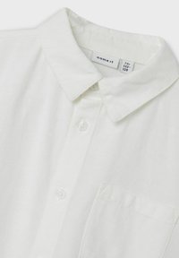 Camicia bianca con bottoni e colletto, realizzata in tessuto testurizzato. Presenta una tasca a sinistra e bottoni bianchi. Etichetta visibile all'interno del colletto.