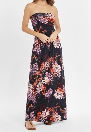 Czarna, długa sukienka bez ramiączek z pomarańczowym, fioletowym i białym kwiatowym wzorem, noszona z płaskimi sandałami, modelka z rękami splecionymi na talii.