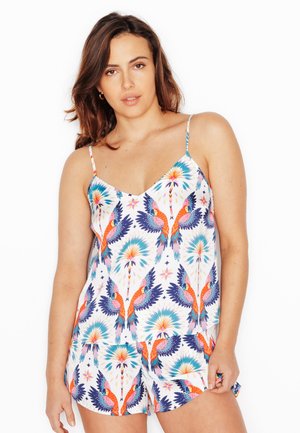 Vrouw draagt kleurrijk mouwloos top met papegaaienprint en bijpassende korte broek, staand tegen een witte achtergrond.