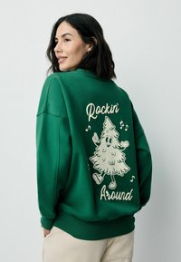 Grøn sweatshirt med en afslappet pasform, der præsenterer en tegneserie-træ grafik og teksten "Rockin' Around" på bagsiden. Blød tekstur.