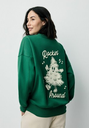 Zielona bluza z luźnym krojem, zdobiona graficznym motywem drzewa w stylu kreskówkowym oraz napisem "Rockin' Around" na plecach. Miła w dotyku tkanina.