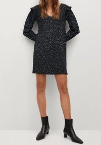 Robe noire à pois avec de longues manches froncées, décolleté en V, longueur au-dessus du genou, associée à des bottines noires. Texture de tissu lisse.