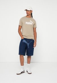 Bež pamukasta majica s bold bijelim "PUMA" logotipom, u kombinaciji s plavim traper šortsevima s grafičkim otisnutim logotipom, bijelim tenisicama i svijetlom kapom.