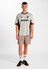 Graues und kastanienbraun gestreiftes Trikot mit kurzen Ärmeln, kombiniert mit beigen Shorts. Das Trikot zeigt ein Logo und Text, getragen mit schwarzen Schuhen und weißen Socken.