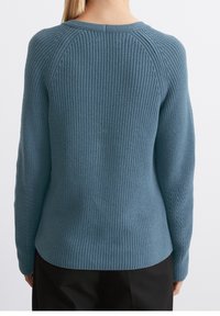 Personne aux longs cheveux blonds portant un pull en tricot côtelé bleu ajusté et un pantalon noir, vue de dos sur un fond uni.