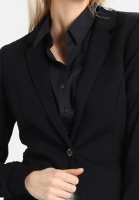 Kvinna som bär en figurnära svart kavaj över en knappad svart skjorta, som justerar kavajen med ena handen vid midjan.