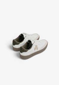 Un par de zapatillas de cuero blanco con bordado de calaveras y huesos cruzados en beige, cuellos de talón de ante y suelas texturizadas en marrón, sobre un fondo blanco.