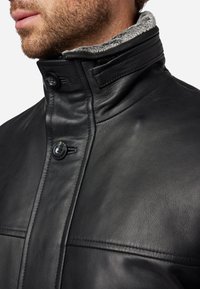 Schwarze Lederjacke mit hohem Kragen, grauem Fellfutter, Knopfverschluss und weicher Textur; sichtbare Nahtdetails.