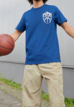 Person iført en blå Puma T-shirt og beige bukser, der holder en basketball udendørs nær en grå væg.