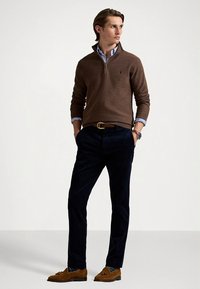 Καφέ πουλόβερ με φερμουάρ στο λαιμό πάνω από ριγέ πουκάμισο με κουμπιά, σετ με σκούρο μπλε παντελόνι και καφέ μοκασίνια. Σημαντικές λεπτομέρειες περιλαμβάνουν τη ζώνη και το ρολόι.