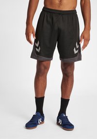 Pantalones cortos atléticos negros hechos de una tela ligera con acentos laterales grises y diseños de rayas en ángulo, combinados con zapatos deportivos azules y calcetines negros.