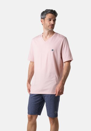 Homme portant un T-shirt rose clair à col en V avec un petit logo sur la poitrine et un short bleu marine à motifs, debout devant un fond uni.