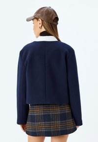 Blazer corto navy con una texture liscia e un collo rotondo, abbinato a una gonna a quadri con tonalità blu, marrone e oro.