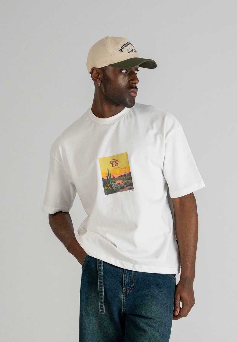 Camiseta blanca de manga corta con un gráfico rectangular colorido que muestra un paisaje desértico. Combinada con una gorra beige y verde.
