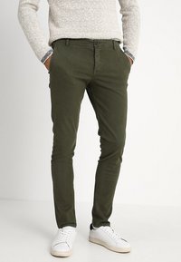 Uomo che indossa pantaloni slim-fit verde oliva, un maglione lavorato a maglia beige e sneakers bianche, in piedi con le mani in tasca.