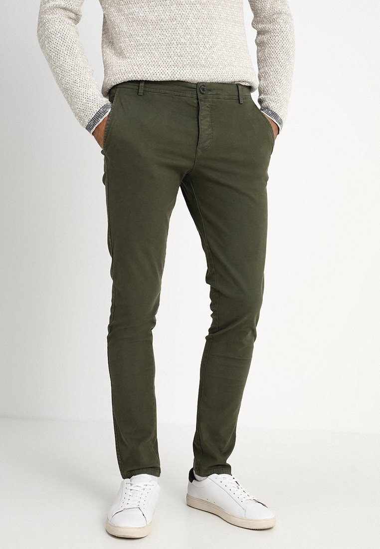 Uomo che indossa pantaloni slim-fit verde oliva, un maglione lavorato a maglia beige e sneakers bianche, in piedi con le mani in tasca.