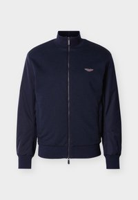 Veste bleu marine à fermeture éclair avec col montant, poignets côtelés et petit logo ailé marron clair sur la poitrine gauche.