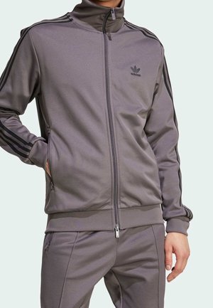 Mężczyzna ubrane w szary dres Adidas z czarnymi paskami oraz kurtką i spodniami na zamek, stojący z jedną ręką w kieszeni.