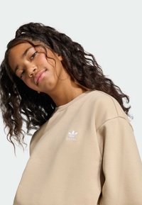 Béžová mikina s bielym vyšívaným logom. Mäkká látka, voľný oversize strih, okrúhly výstrih a spustené ramená. Kučeravé vlasy rámujúce tvár.