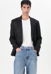 Blazer negro con un solo botón, llevado sobre una camiseta blanca. Combinado con jeans ligeros de corte holgado en color azul. Tejido suave, ajuste entallado.