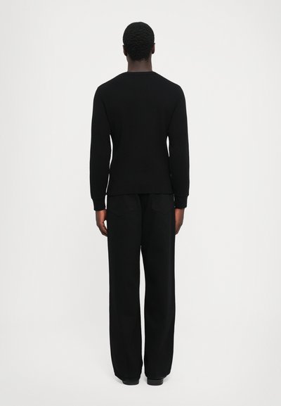 Vīrietis stāv ar muguru pret kameru, ģērbies melnā garām piedurknēm kreklā un brīvās melnās biksēs uz vienkrāsaina baltā fona.