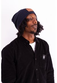 Navy knitted beanie with a textured finish and a brown leather patch. Black corduroy button-up shirt with logo on the chest pocket.