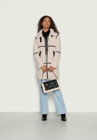 Casaco de fleece bege com acabamentos e fechos pretos, combinado com calças de ganga azul claro e uma mala preta acolchoada. Textura suave, design casual.