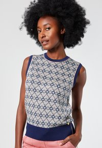 Sleeveless knit sweater features a geometric pattern in pale pink and navy on a light background, with a ribbed navy hem and side button detail.